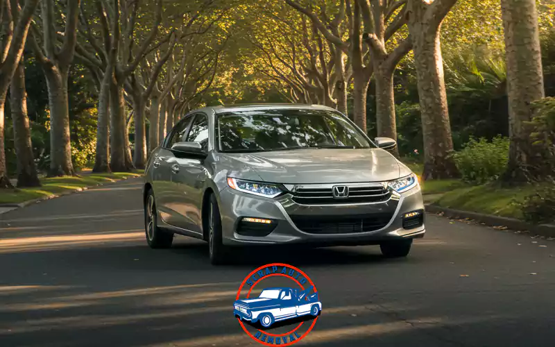 Silver car on a tree-lined street. The car is a Honda. A red and blue logo is at the bottom.