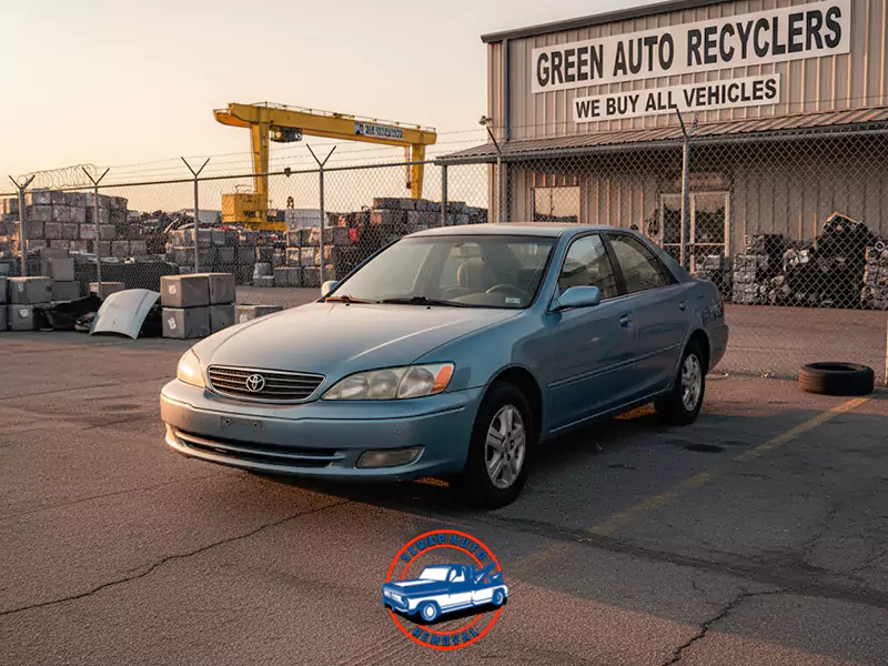 Used blue sedan at "Green Auto Recyclers" facility at sunset. Scrap metal and crane visible.
