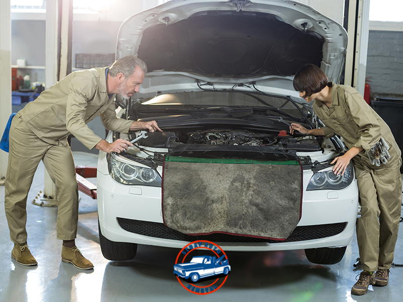 Two mechanics inspecting the engine of a white car with tools and protective mats in a bright auto shop