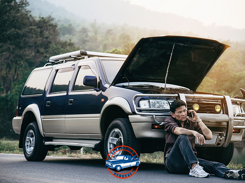 Man sitting beside a broken-down SUV with the hood open, making a phone call on a roadside near the woods