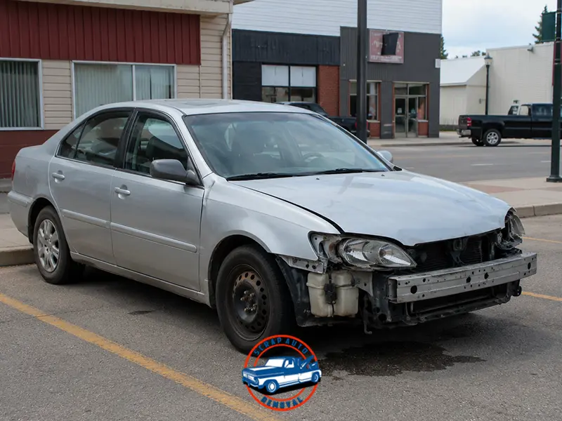 Silver sedan missing front bumper and headlight, parked in a town commercial parking lot.